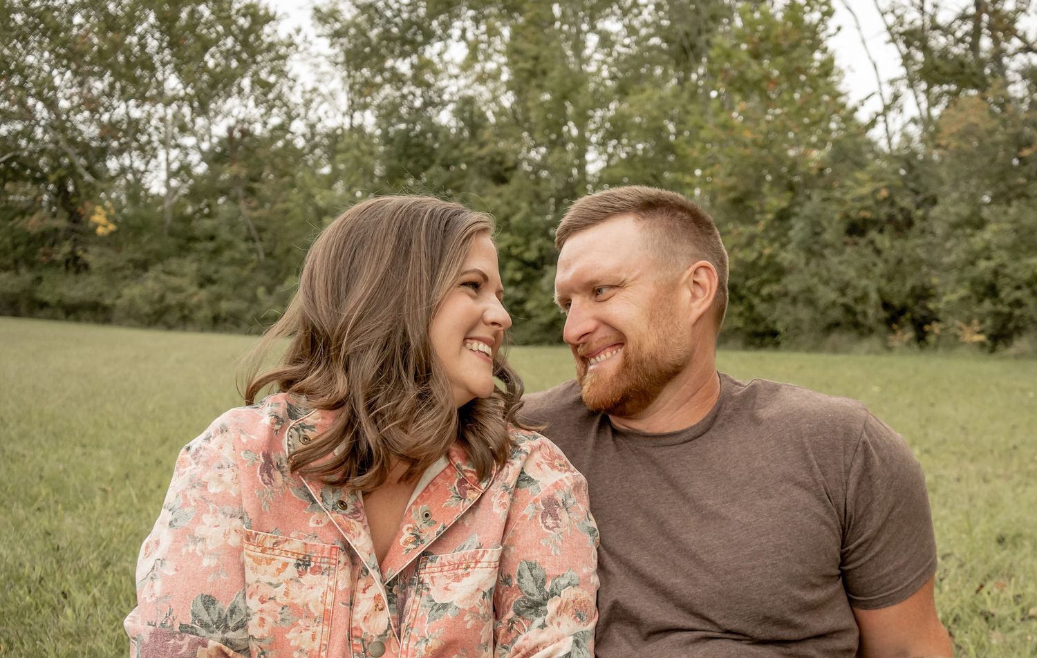Smiling couple sitting together in a lush green field, sharing a joyful moment outdoors.