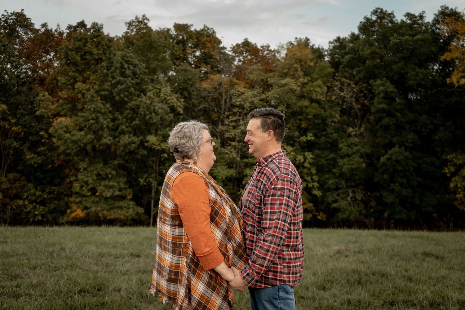 A mature couple holds hands and smiles at each other in a green field with autumn trees behind them.