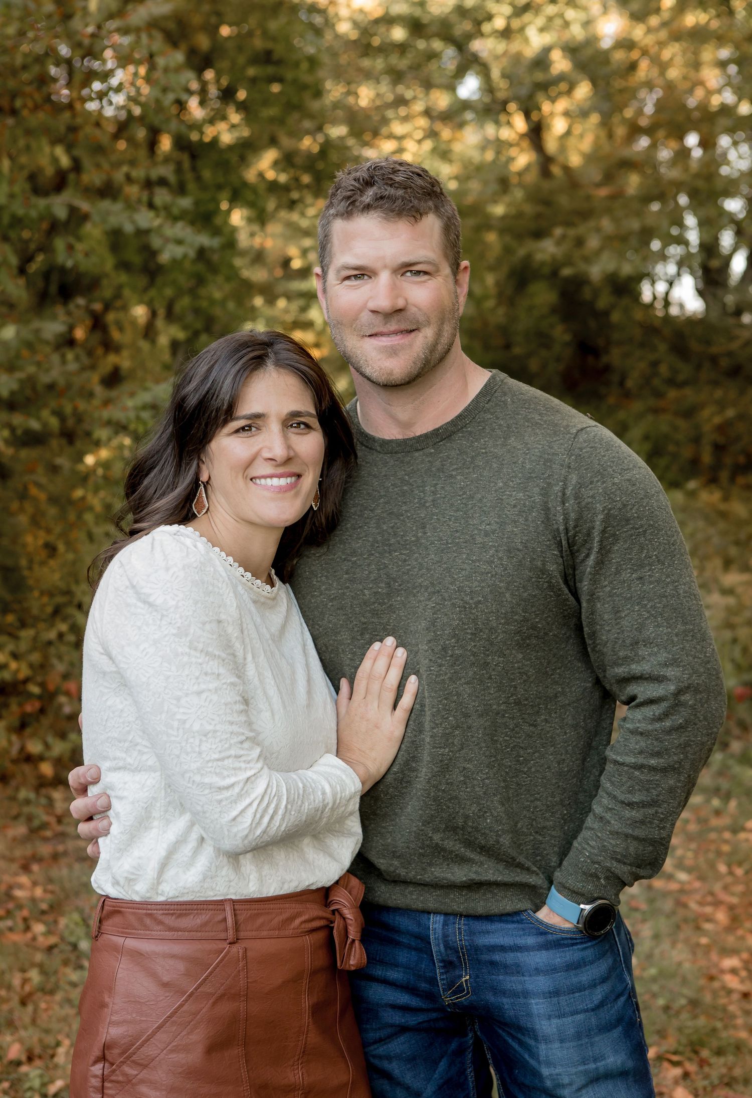 Smiling couple posing together outdoors in autumn, woman in white top and brown skirt, man in green sweater.