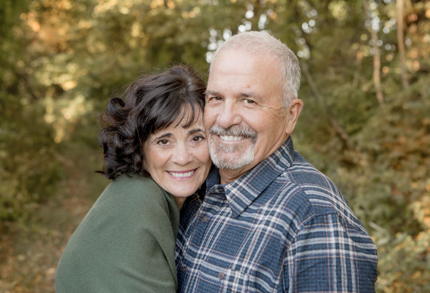 Smiling older couple embracing warmly outdoors among autumn foliage in a wooded setting.