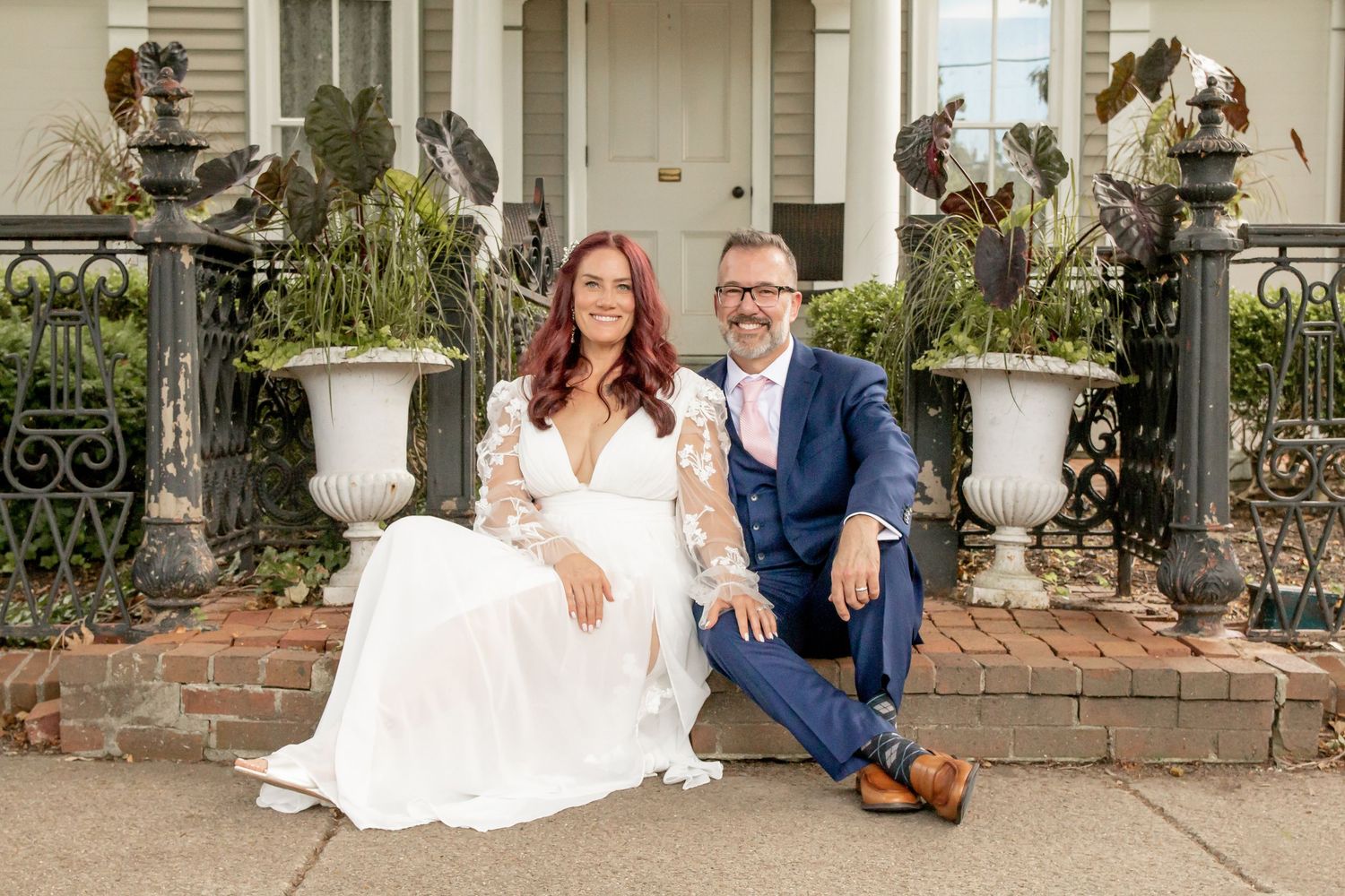 Couple sitting together on brick steps outside elegant building, woman in white wedding dress, man in blue suit.