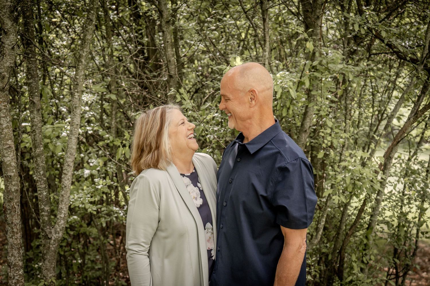Couple smiling at each other surrounded by lush green trees in an outdoor portrait session.