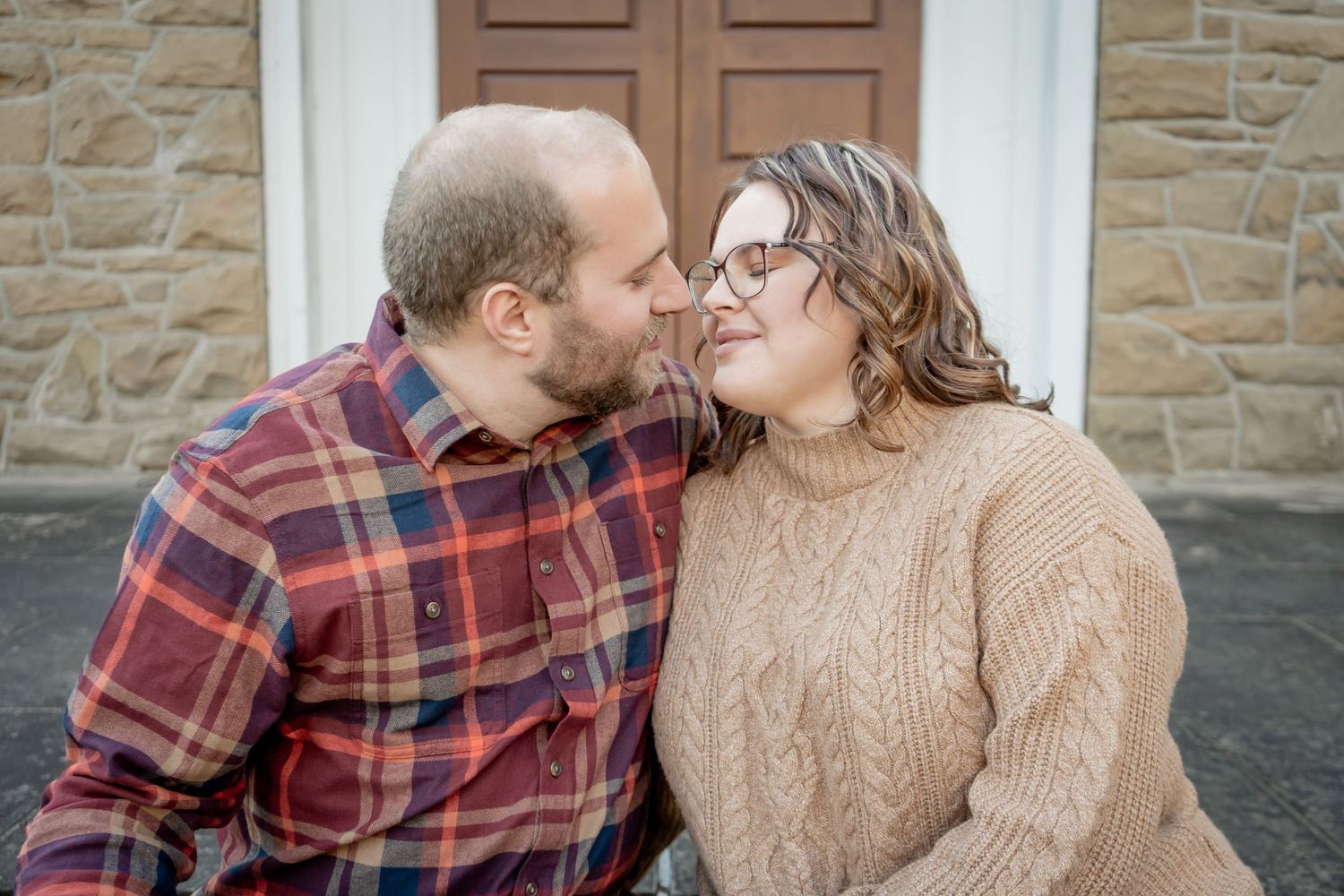 A bearded man and a woman in a knit sweater share a romantic nose-to-nose moment outdoors.