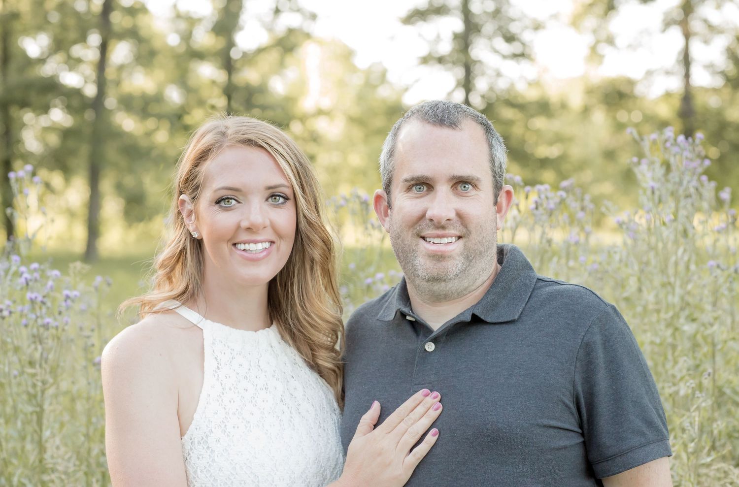 Happy couple posing together in a sunny meadow with wildflowers, woman wearing white dress.