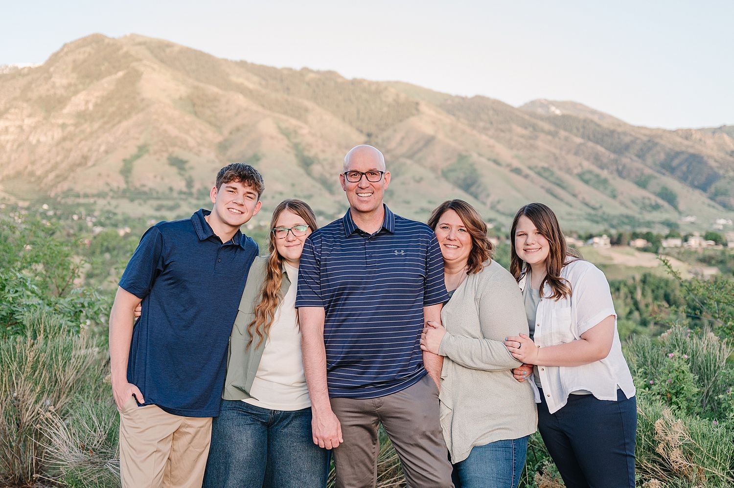 A group of five people stands together outdoors against a backdrop of scenic mountain peaks and green foliage.