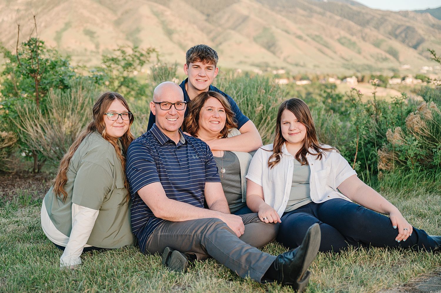 A family group sits together on grass with scenic desert mountains in the background during golden hour.