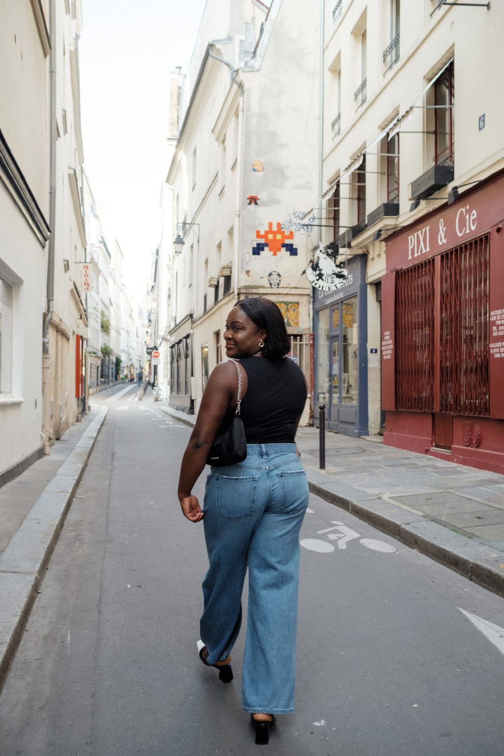 Person walking down a narrow cobblestone street in historic Parisian neighborhood wearing jeans and black top.