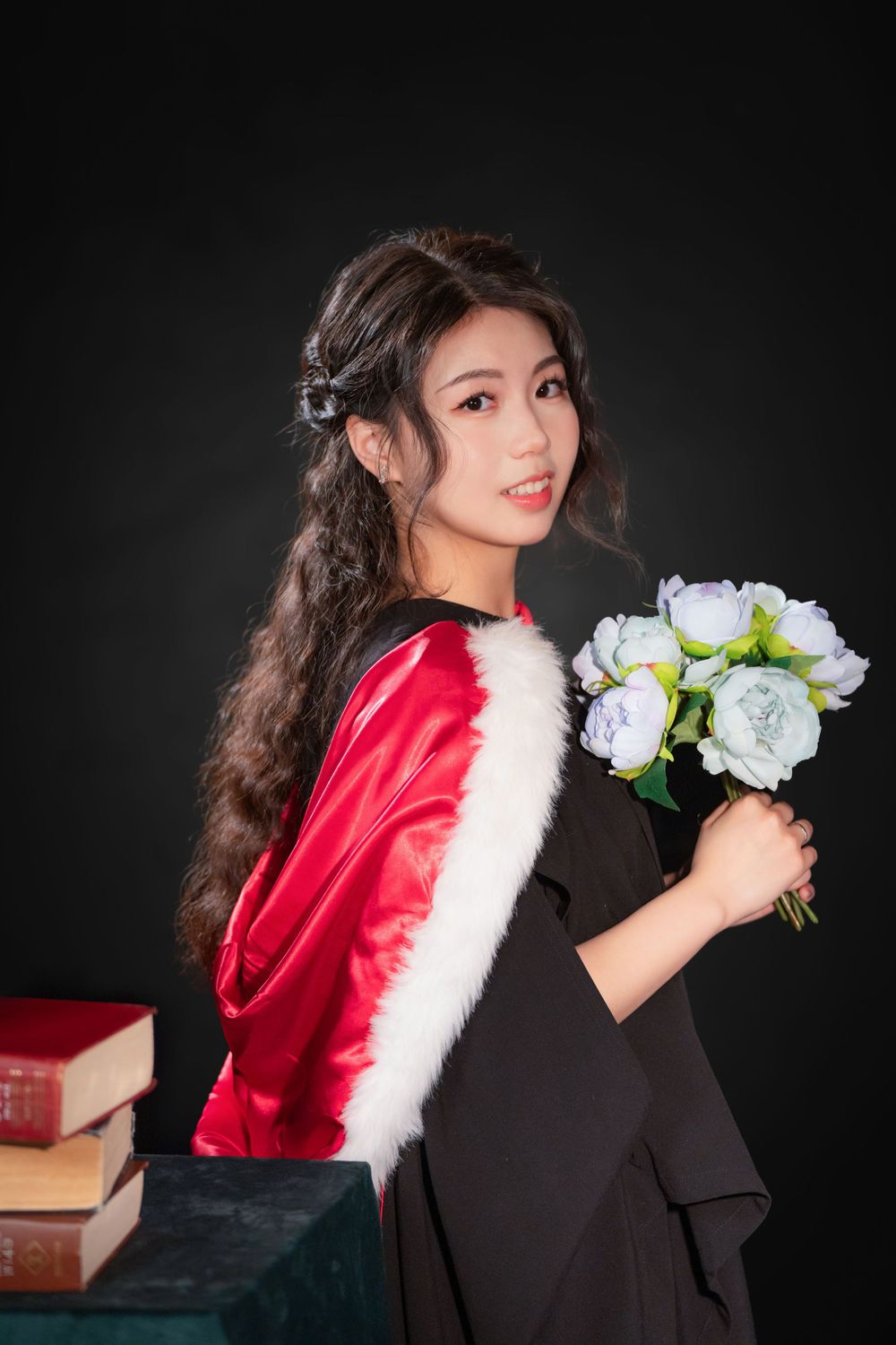 Close-up portrait of a woman in a black graduation gown holding white flowers and books.