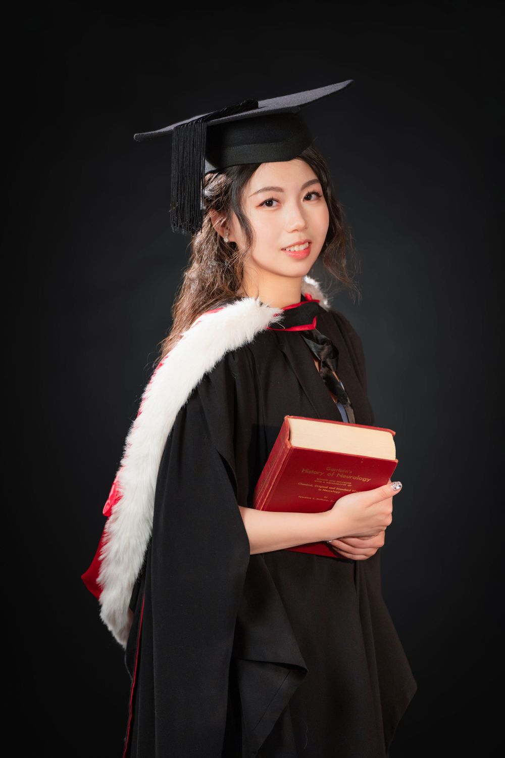 Close-up portrait of a woman in a black graduation gown holding white flowers and books.