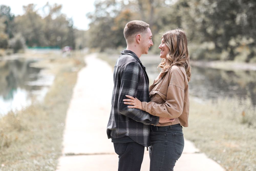 A couple embraces tenderly on a pathway with a peaceful lake and trees in the background.