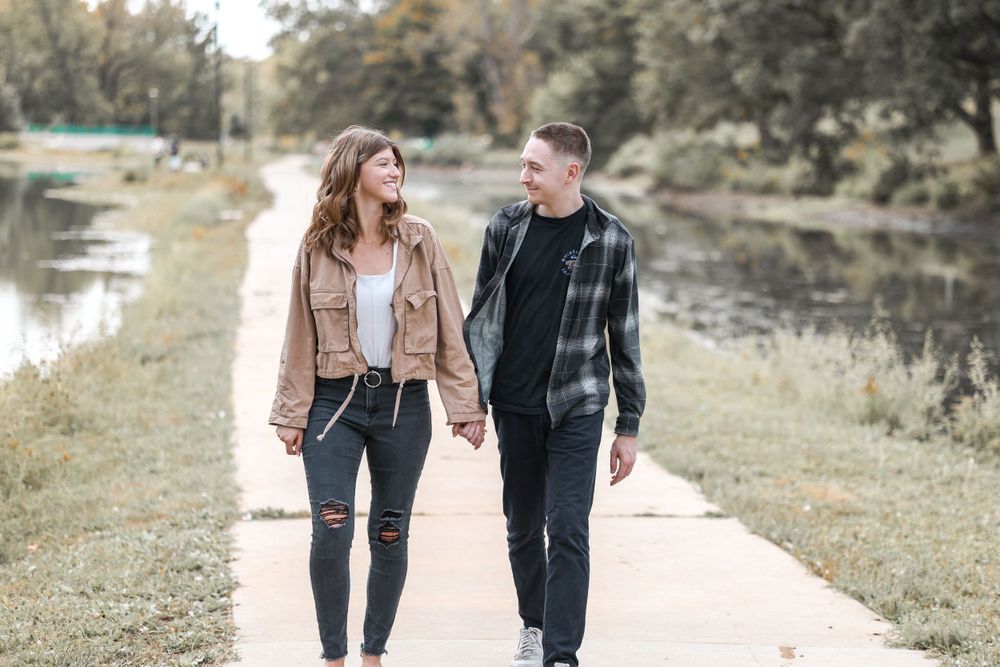 Couple walks hand in hand along a lakeside path wearing casual fall outfits.