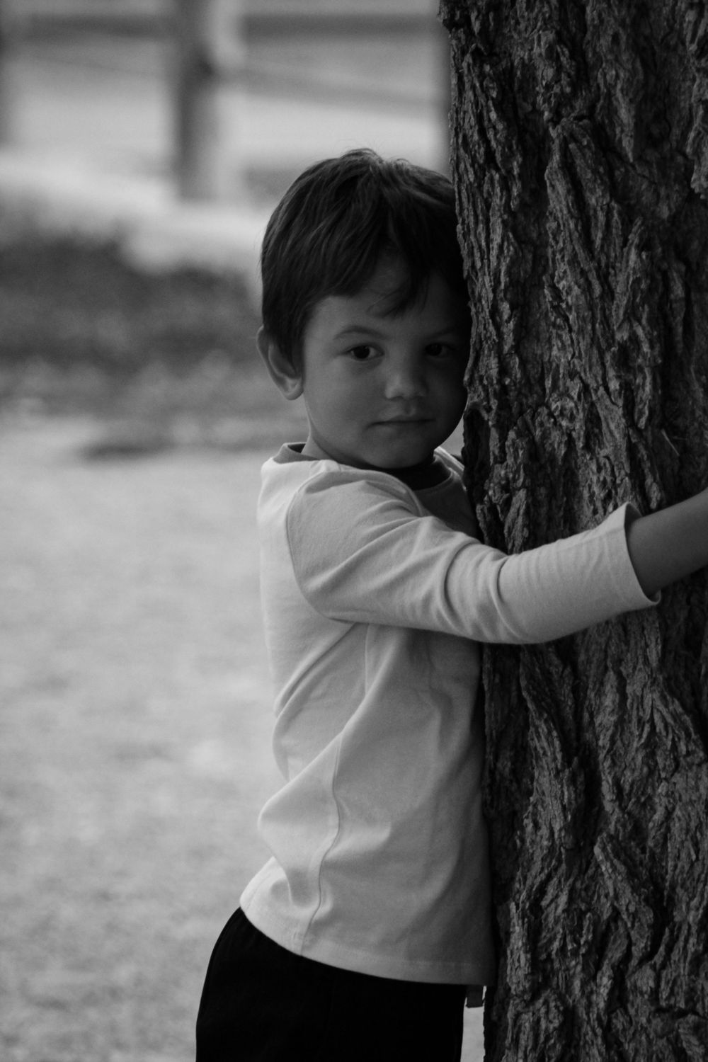 Child in white long sleeve shirt hugging large tree trunk in black and white photo.