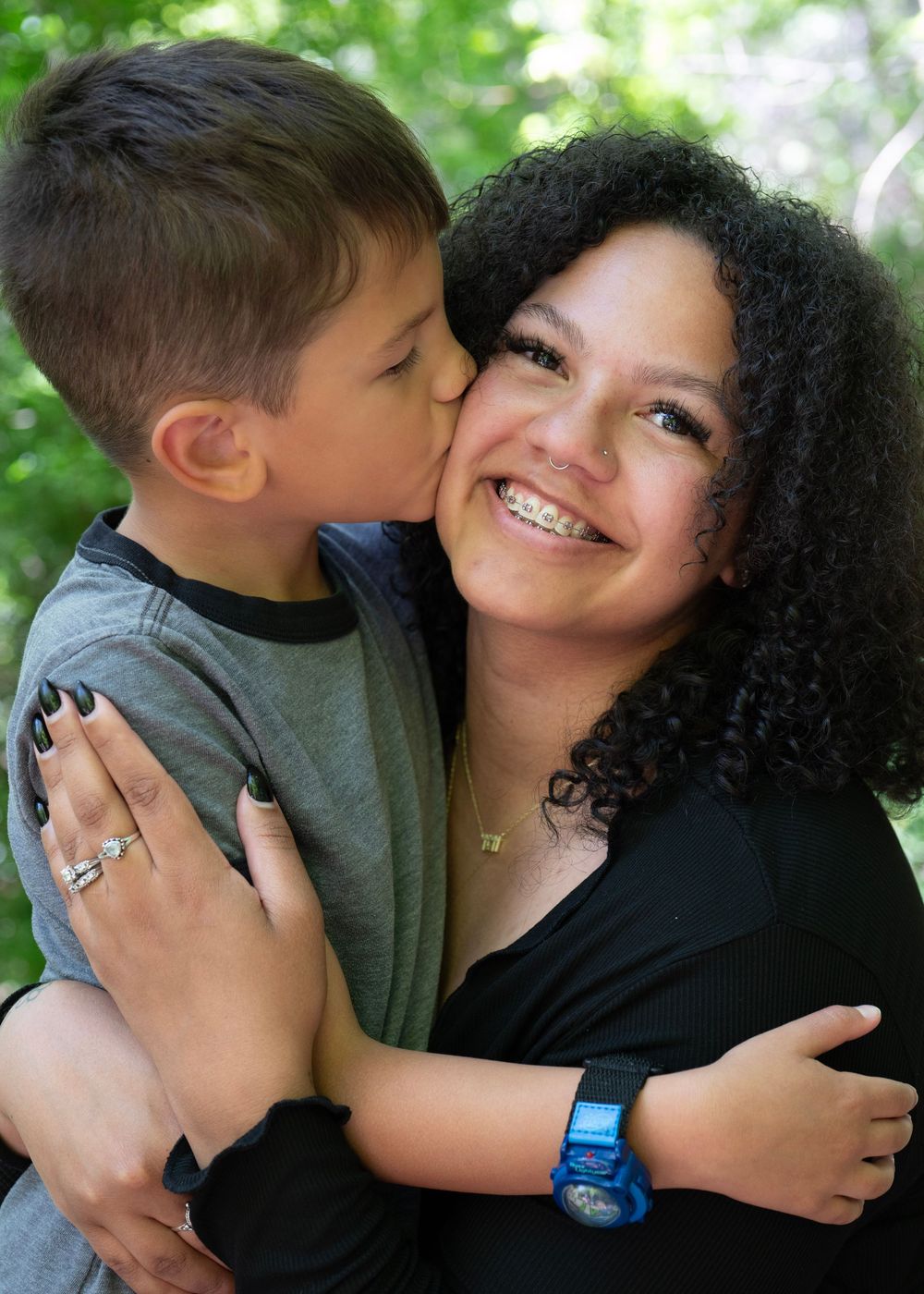 A young boy kisses his smiling mother on the cheek while they embrace outdoors among green trees.