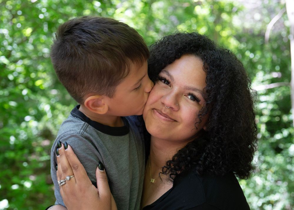 A young boy kisses a curly-haired woman on the cheek outdoors, surrounded by green foliage.