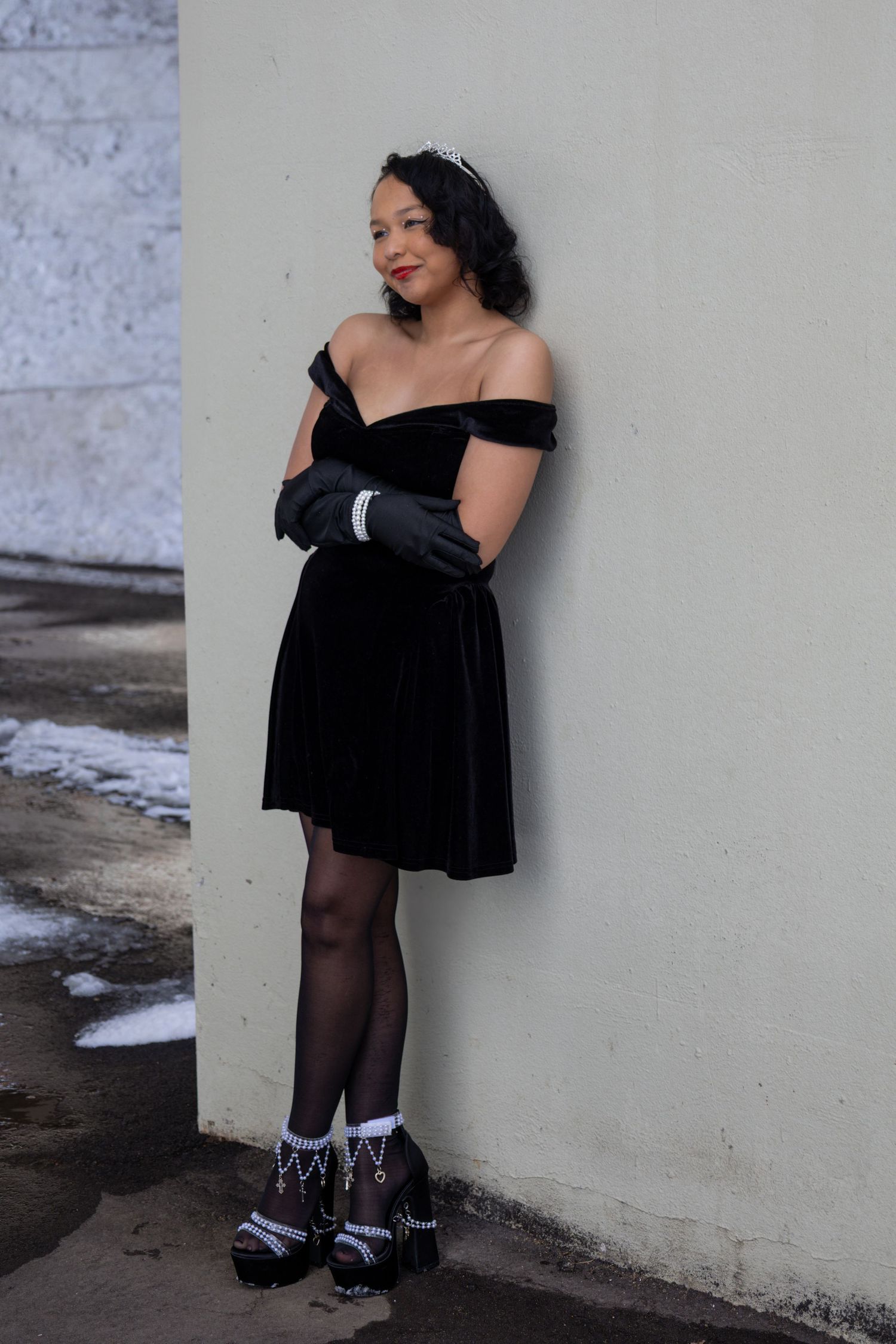 Woman in black dress and gloves poses elegantly against wall.
