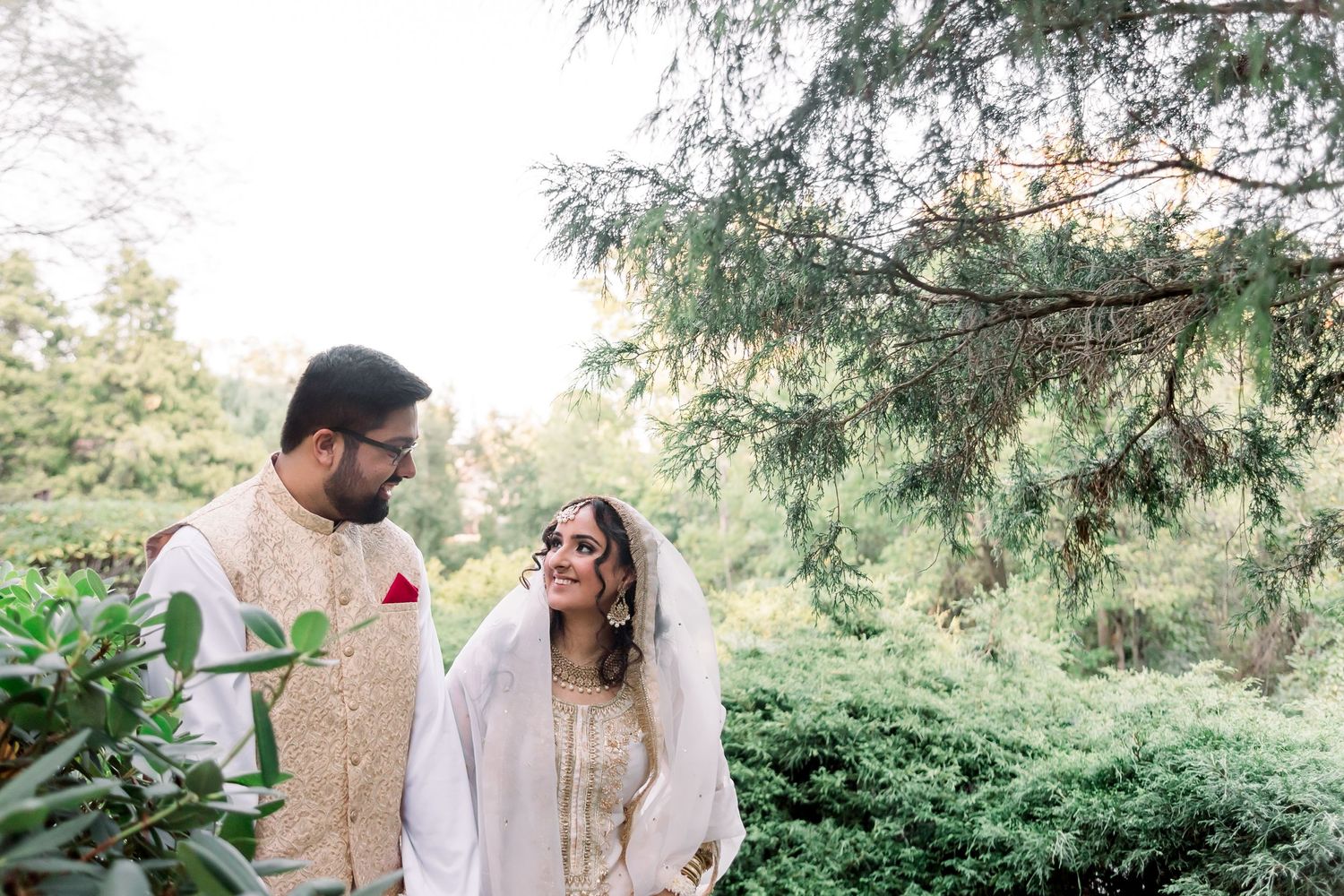 A couple walks together in traditional South Asian wedding attire through a lush garden setting.