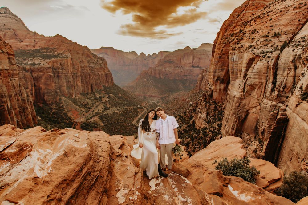 Couple stands on red rock overlook with vast canyon vista in golden light.