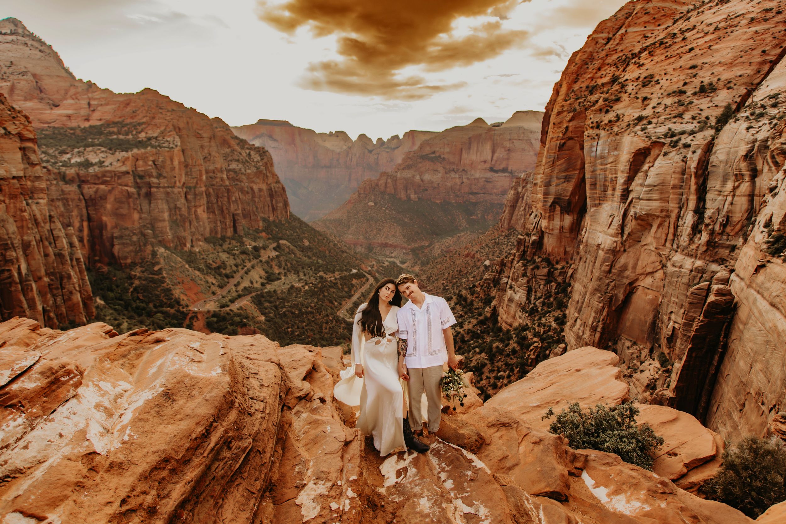 Couple stands on red rock overlook with vast canyon vista in golden light.