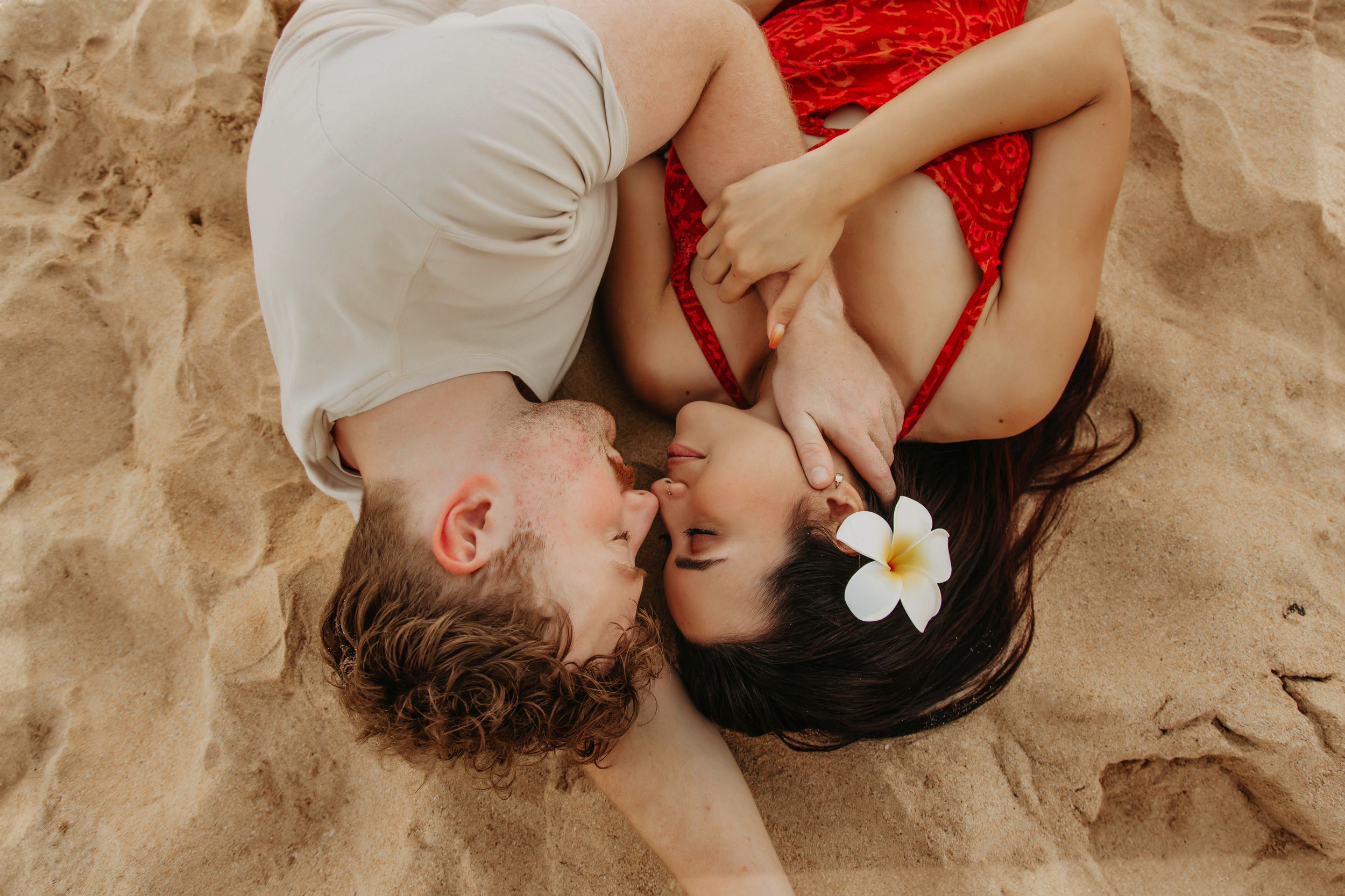 Couple lies on sandy beach with tropical flower detail in intimate pose.
