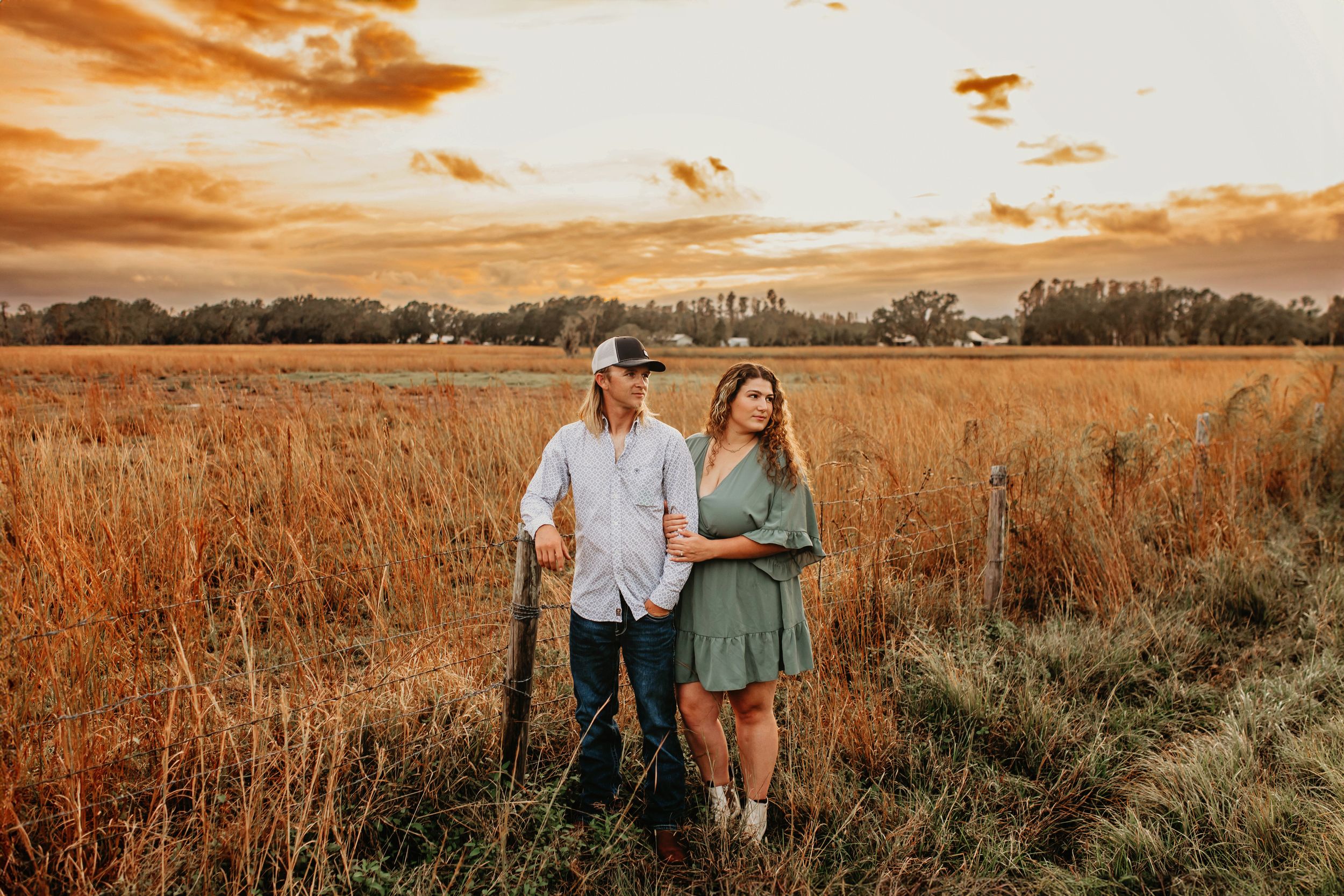 A couple stands in a golden field at sunset with a dramatic orange sky behind them.