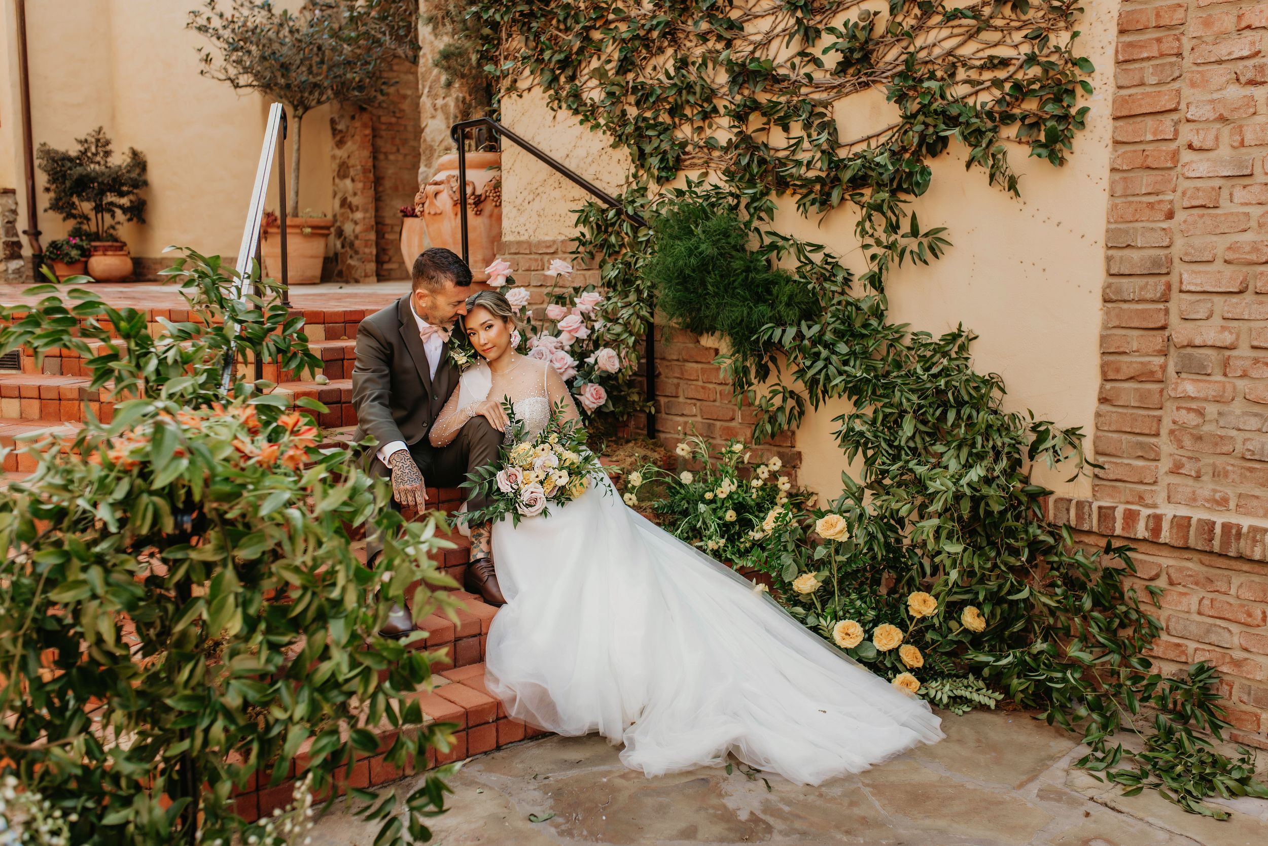 A romantic wedding couple shares an intimate moment on brick steps surrounded by gardens.