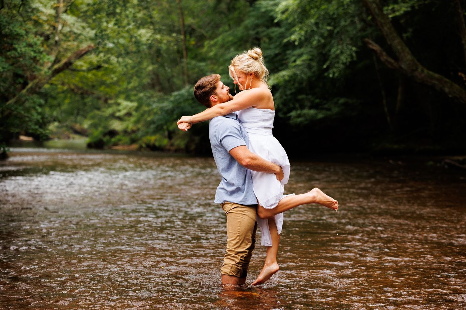 Couple embracing romantically in a shallow forest river, man lifting woman in white dress, lush green trees background.