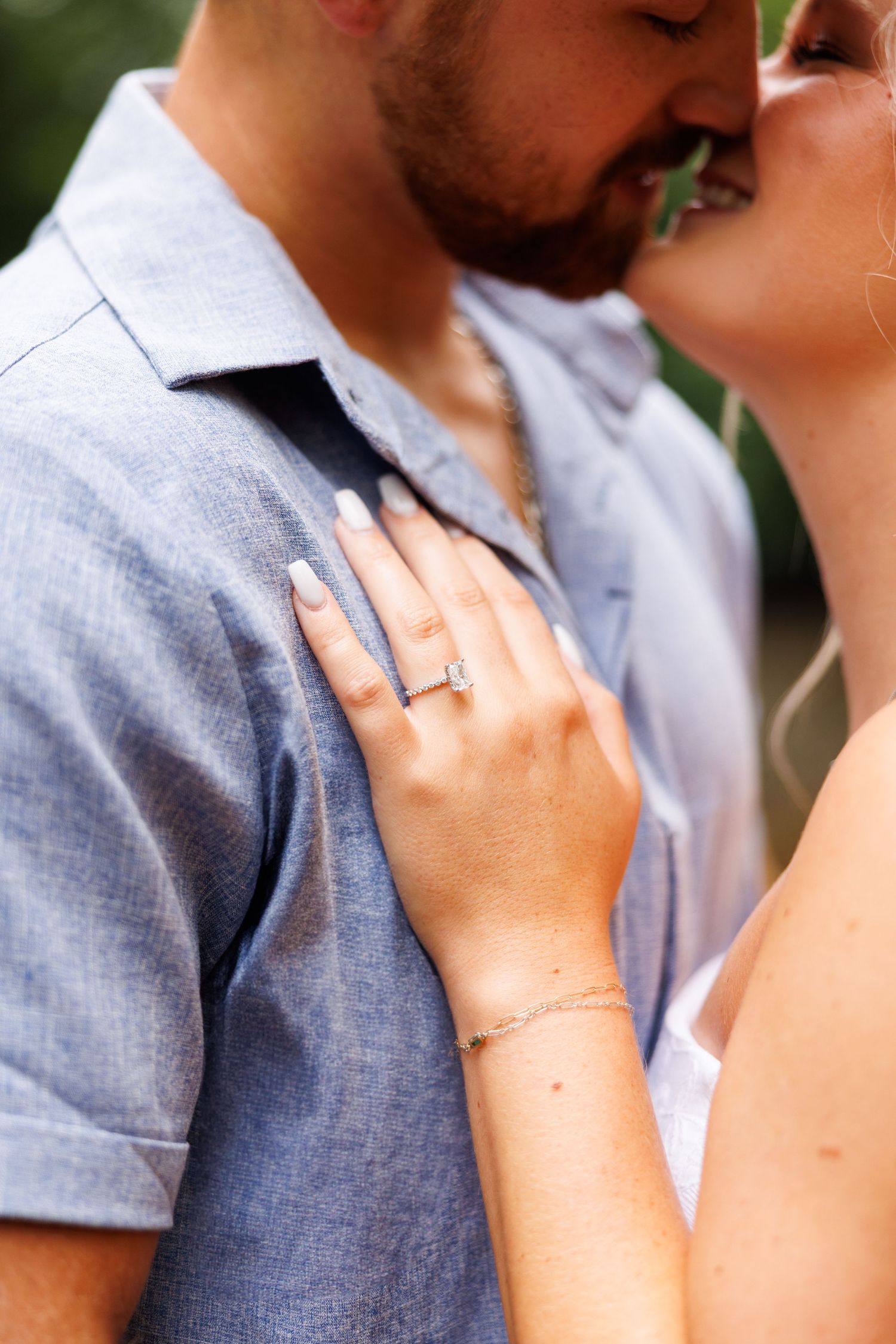 Couple sharing a kiss during engagement photos, woman displaying diamond ring on man's chest.