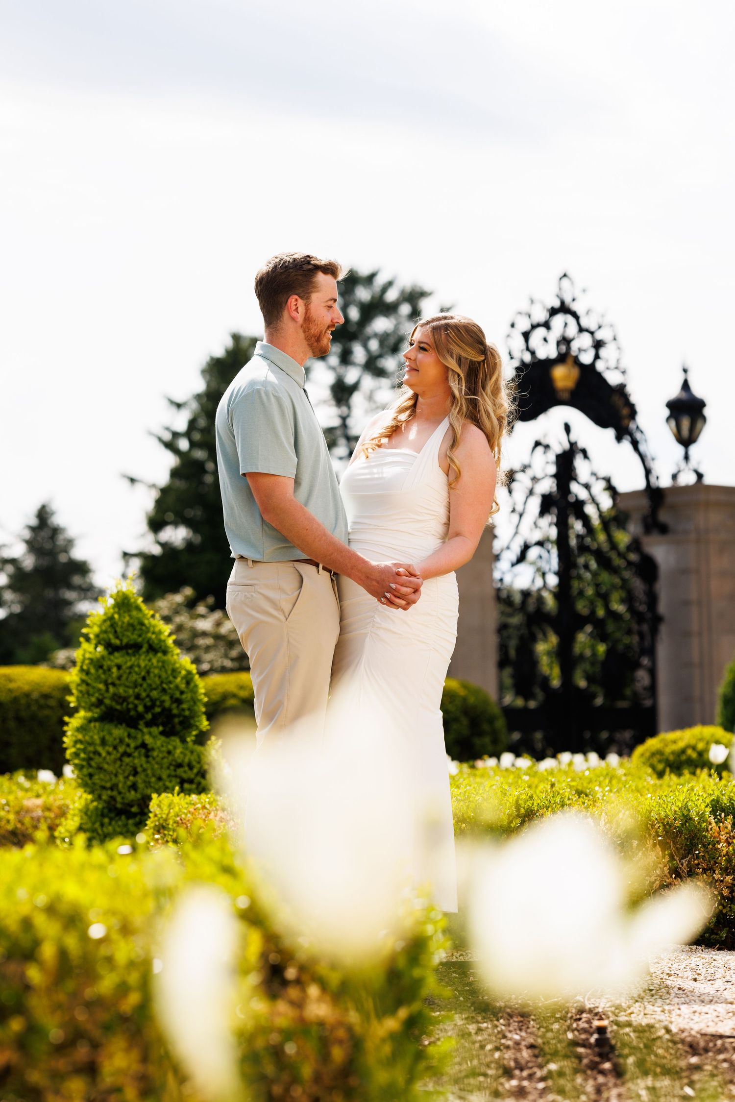 Couple holding hands and smiling at each other in a lush garden with ornate iron gates.