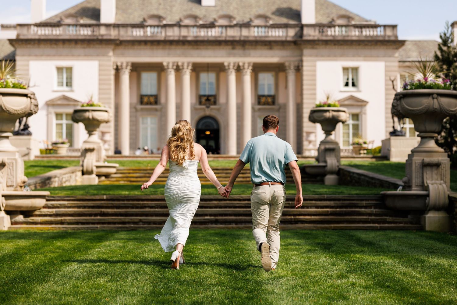 Couple holding hands running toward grand estate mansion with stone steps and manicured lawn.