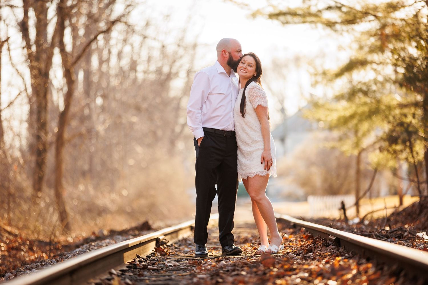 Romantic couple standing on railroad tracks surrounded by autumn trees, man kissing woman's forehead in tender embrace.