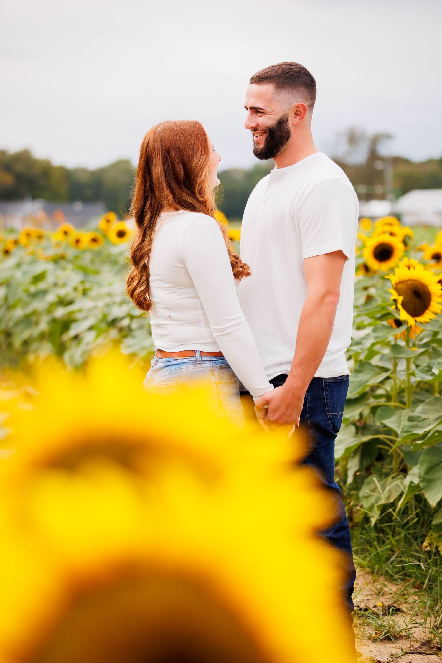 Couple holding hands in a sunflower field, smiling at each other, wearing white tops and jeans on a cloudy day.