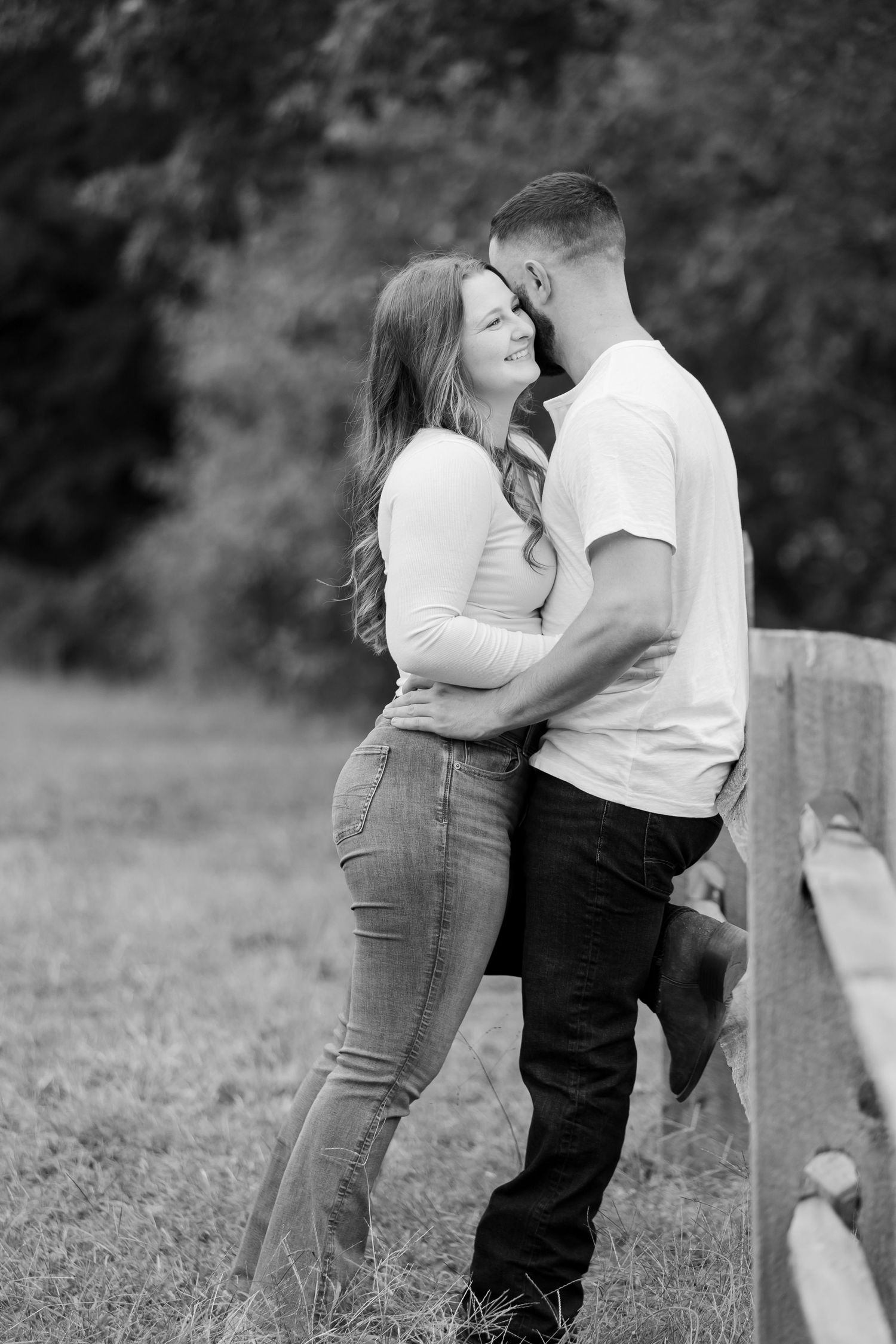 Black and white photo of a romantic couple embracing near a wooden fence in a lush outdoor setting.