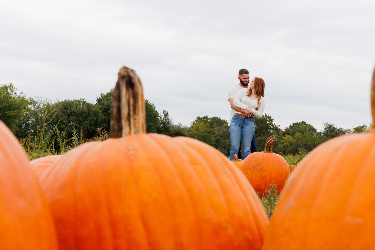 Couple embracing romantically in a pumpkin patch surrounded by large orange pumpkins on a cloudy autumn day.