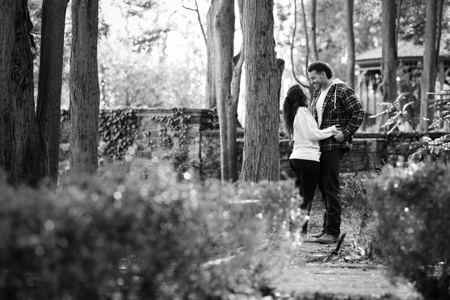 Black and white photo of a romantic couple sharing a tender moment on a garden path surrounded by trees.