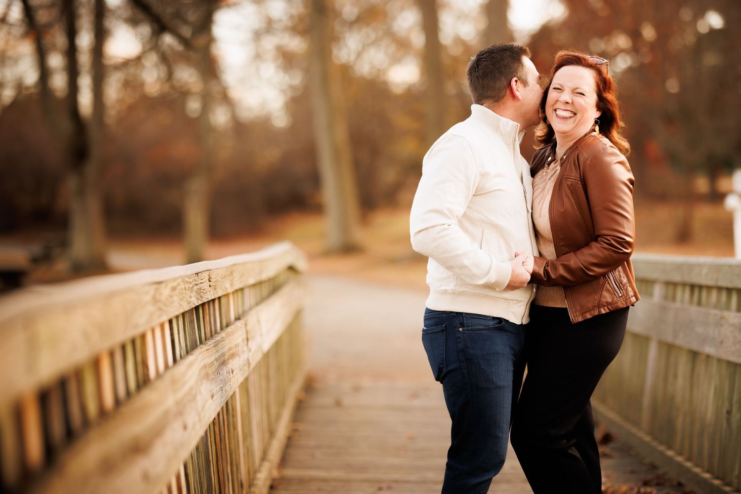Couple sharing a romantic moment on a wooden bridge in autumn park, woman laughing as man kisses her cheek.