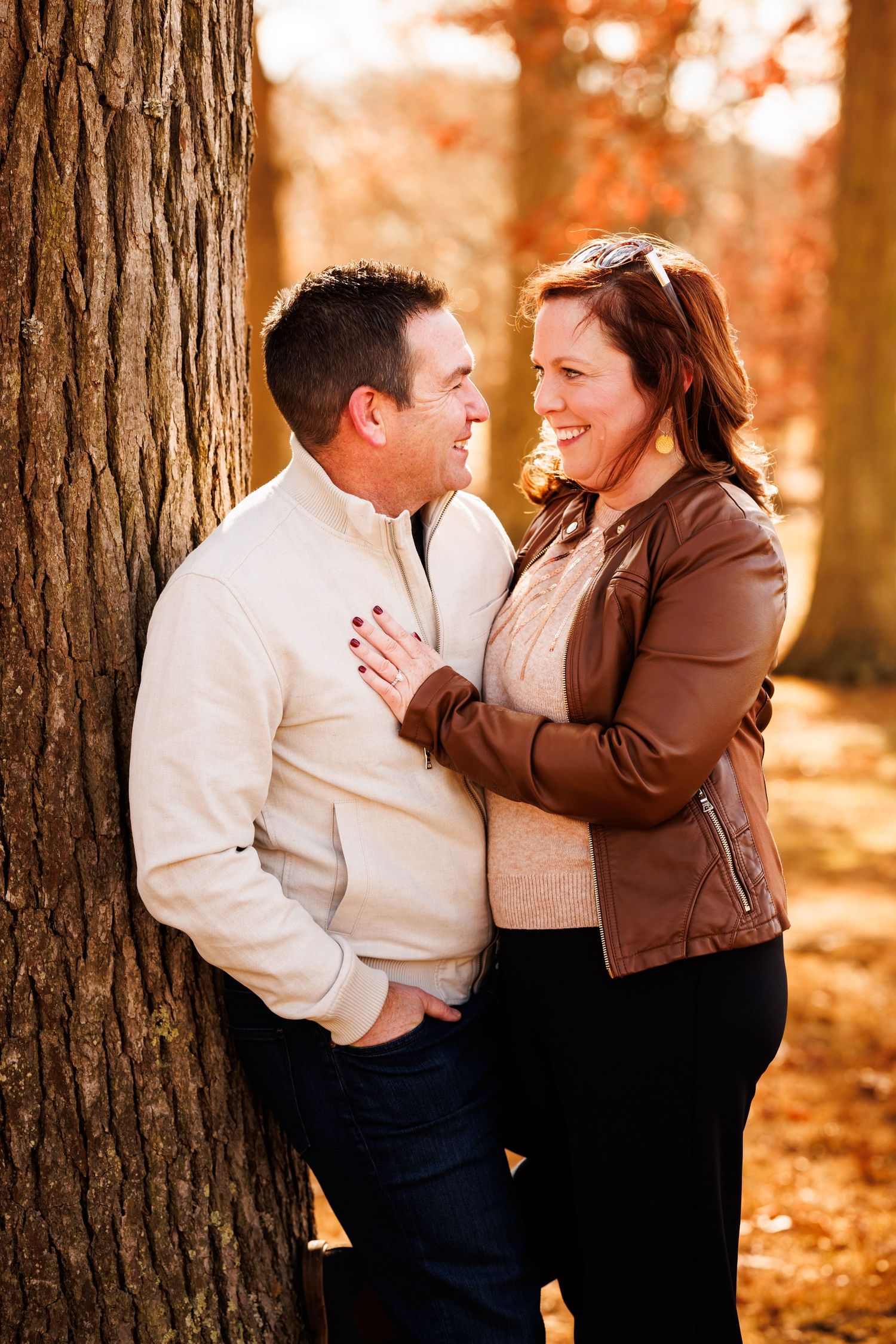 Couple laughing together leaning against a tree in autumn forest, woman in brown leather jacket touching man's chest.
