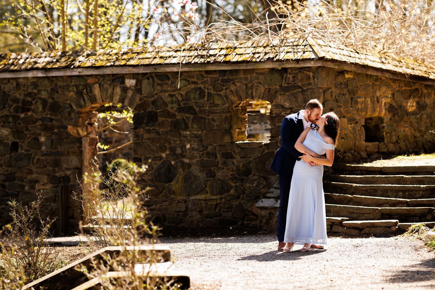 Couple sharing a romantic embrace near a rustic stone building with steps in a sunlit outdoor setting.