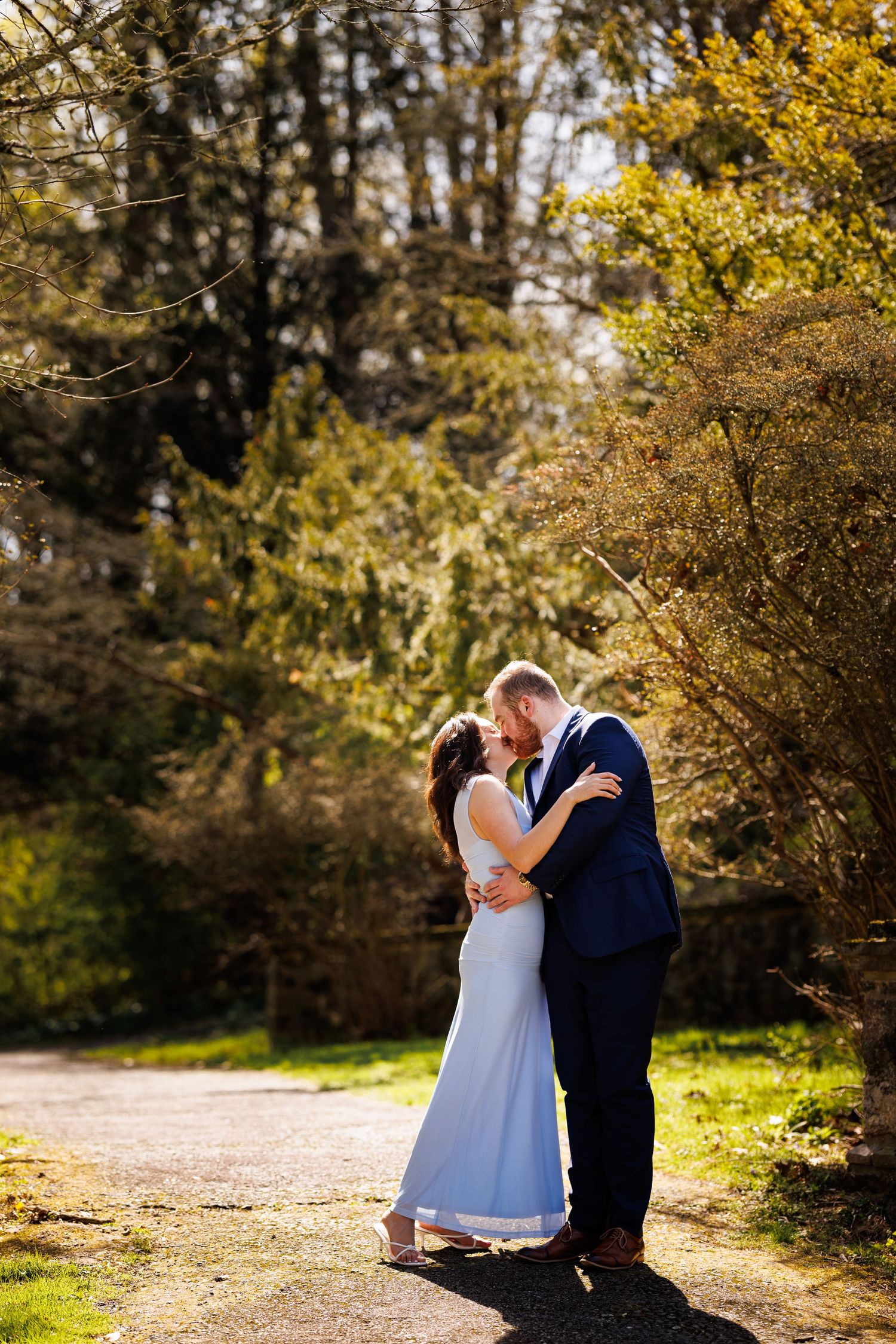 A couple shares a romantic kiss on a sunlit garden path, the bride in a white gown and groom in a navy suit.