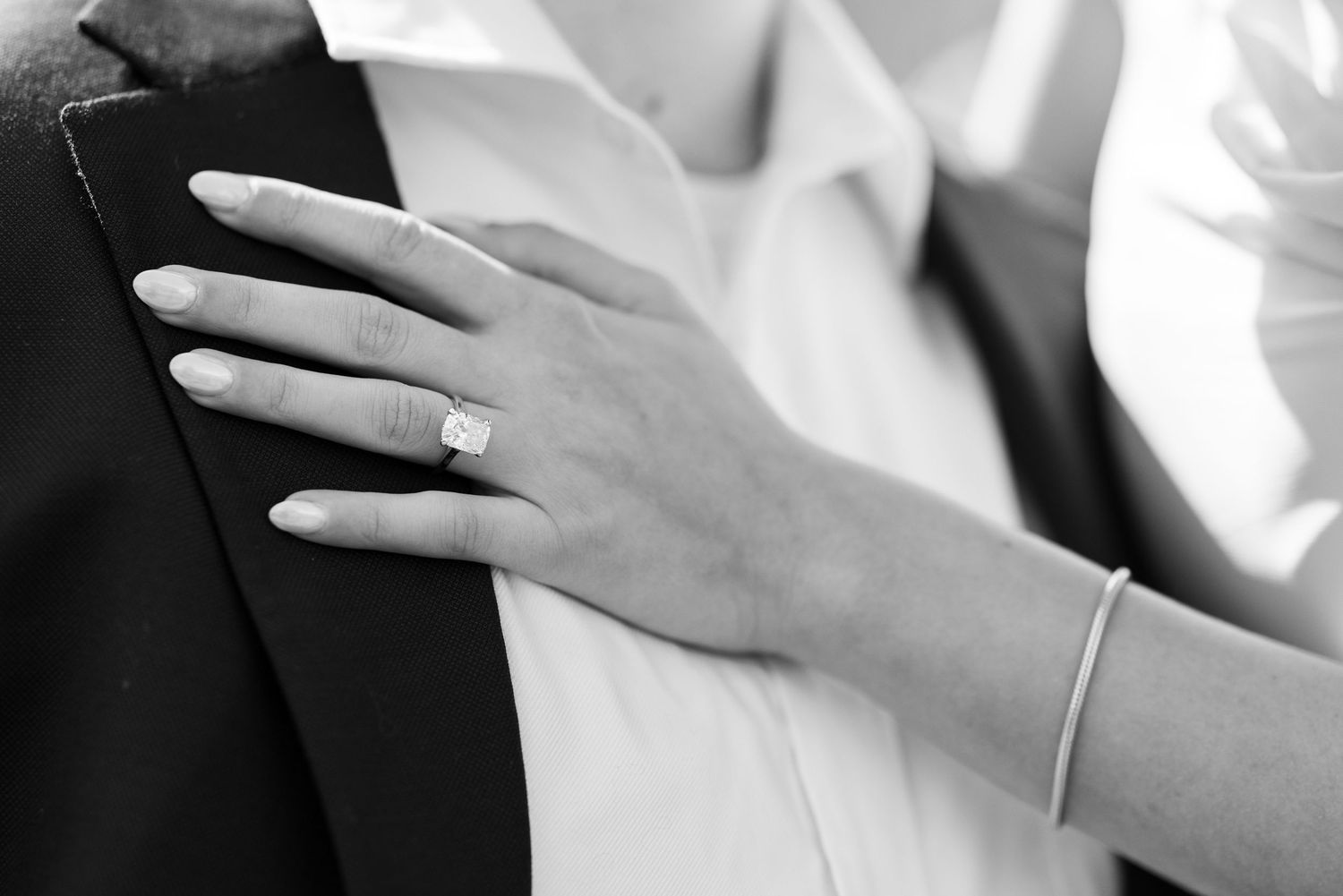 Black and white close-up of a woman's hand with an engagement ring resting on a man's suit jacket.