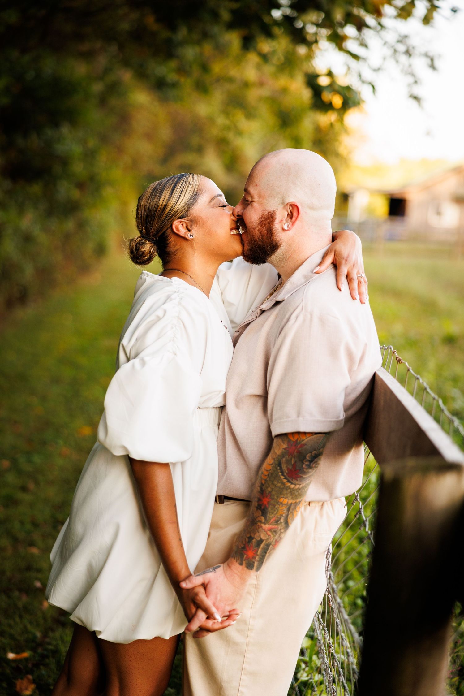 Couple sharing a romantic kiss outdoors near a fence, woman in white dress and tattooed man in beige shirt.
