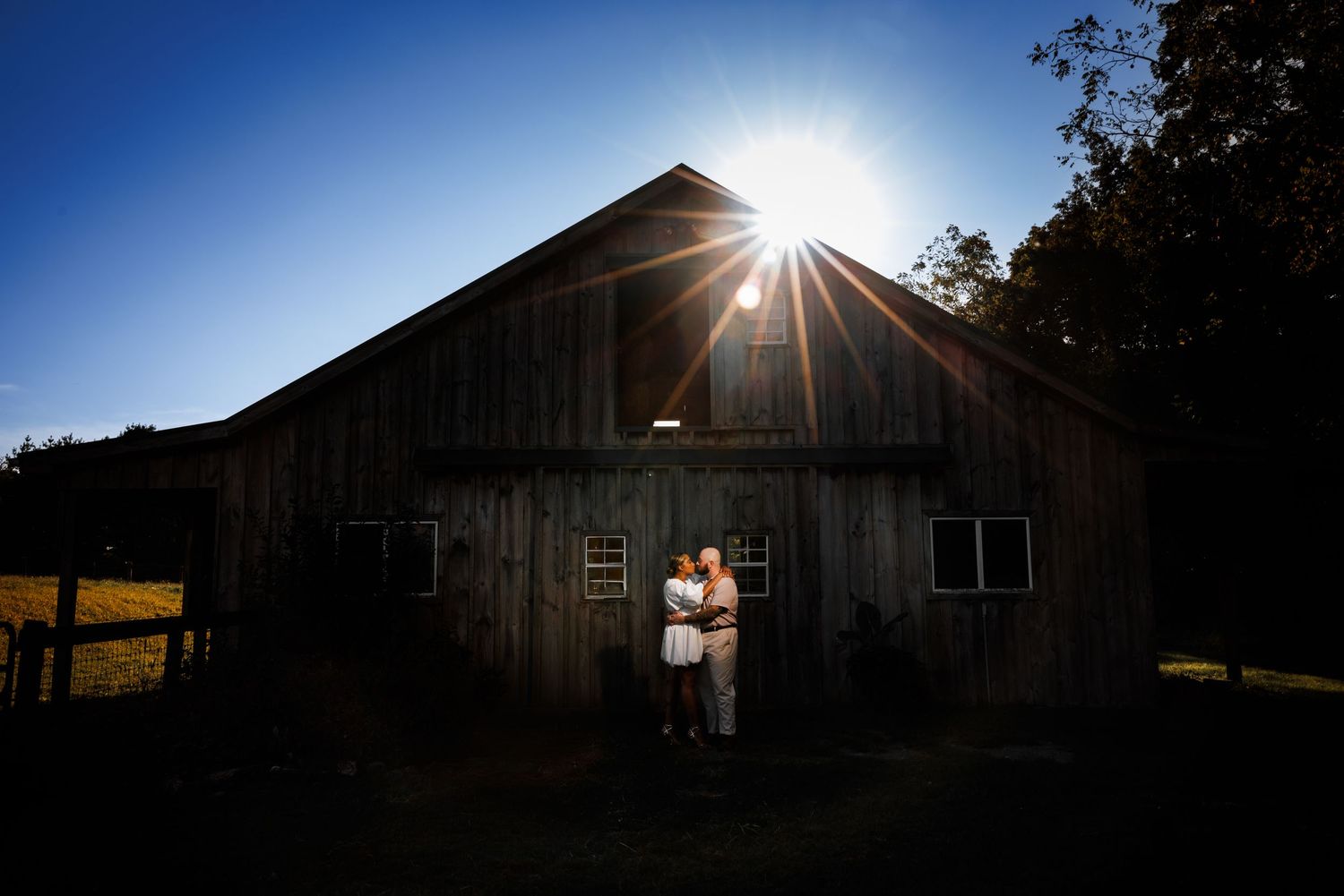 Couple embracing in front of rustic wooden barn at sunset with dramatic sunburst and deep blue sky.