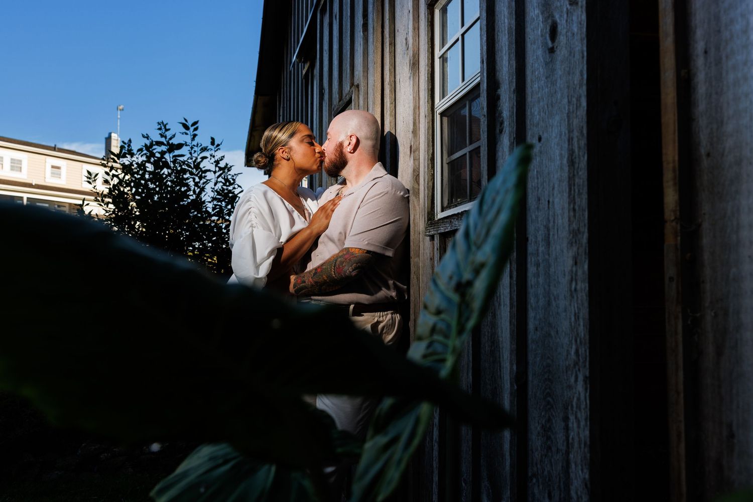 Couple sharing a romantic moment kissing outdoors near a rustic wooden building with soft golden light.