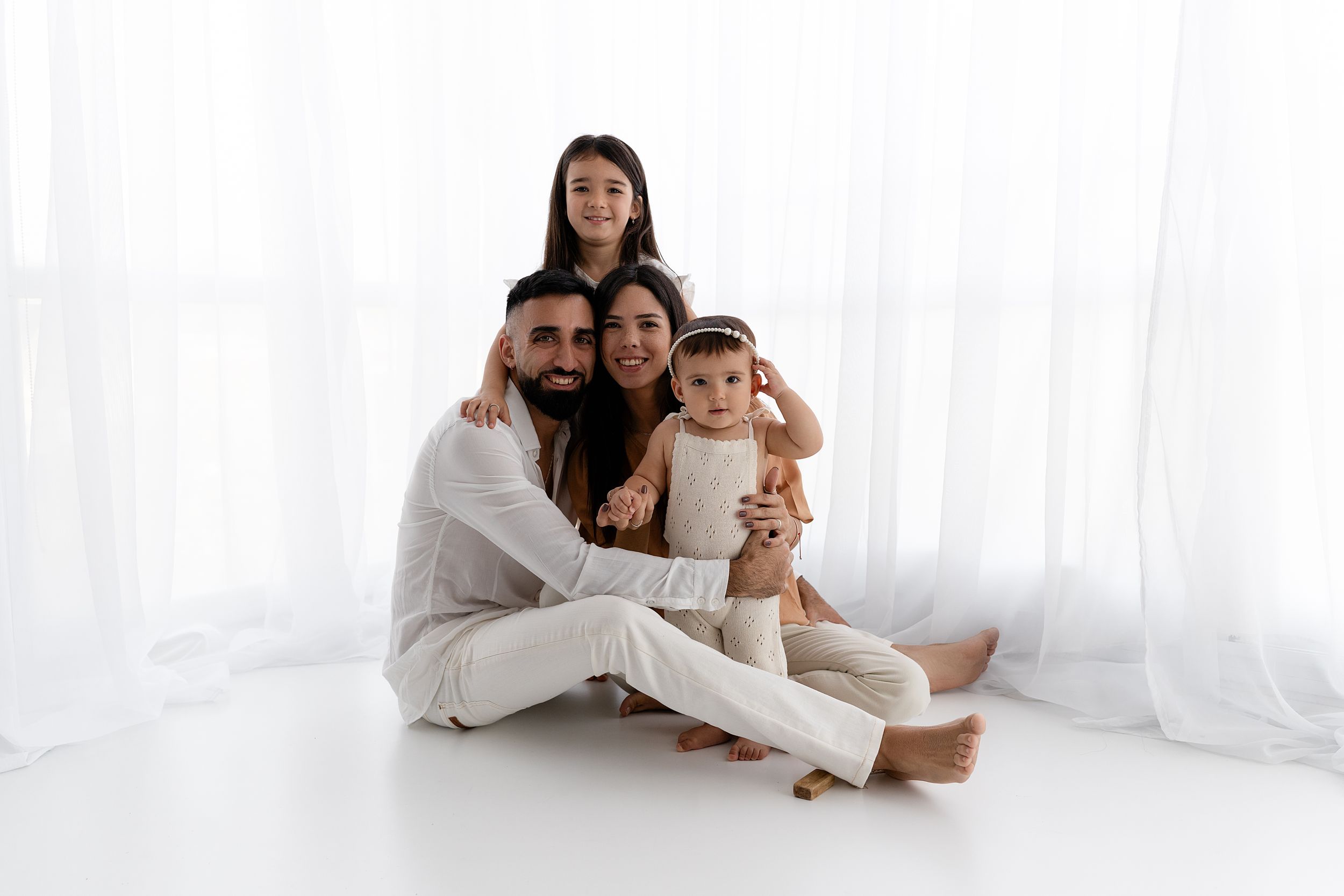 Family wearing coordinated white outfits poses together on white studio floor.