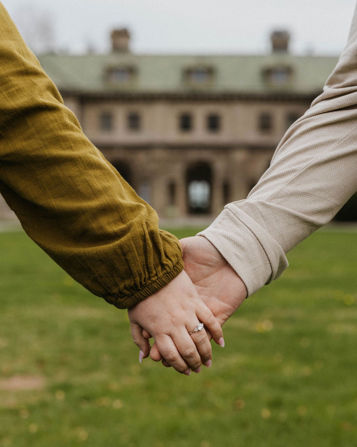 Couple holding hands showing engagement ring with historic stone building in background.