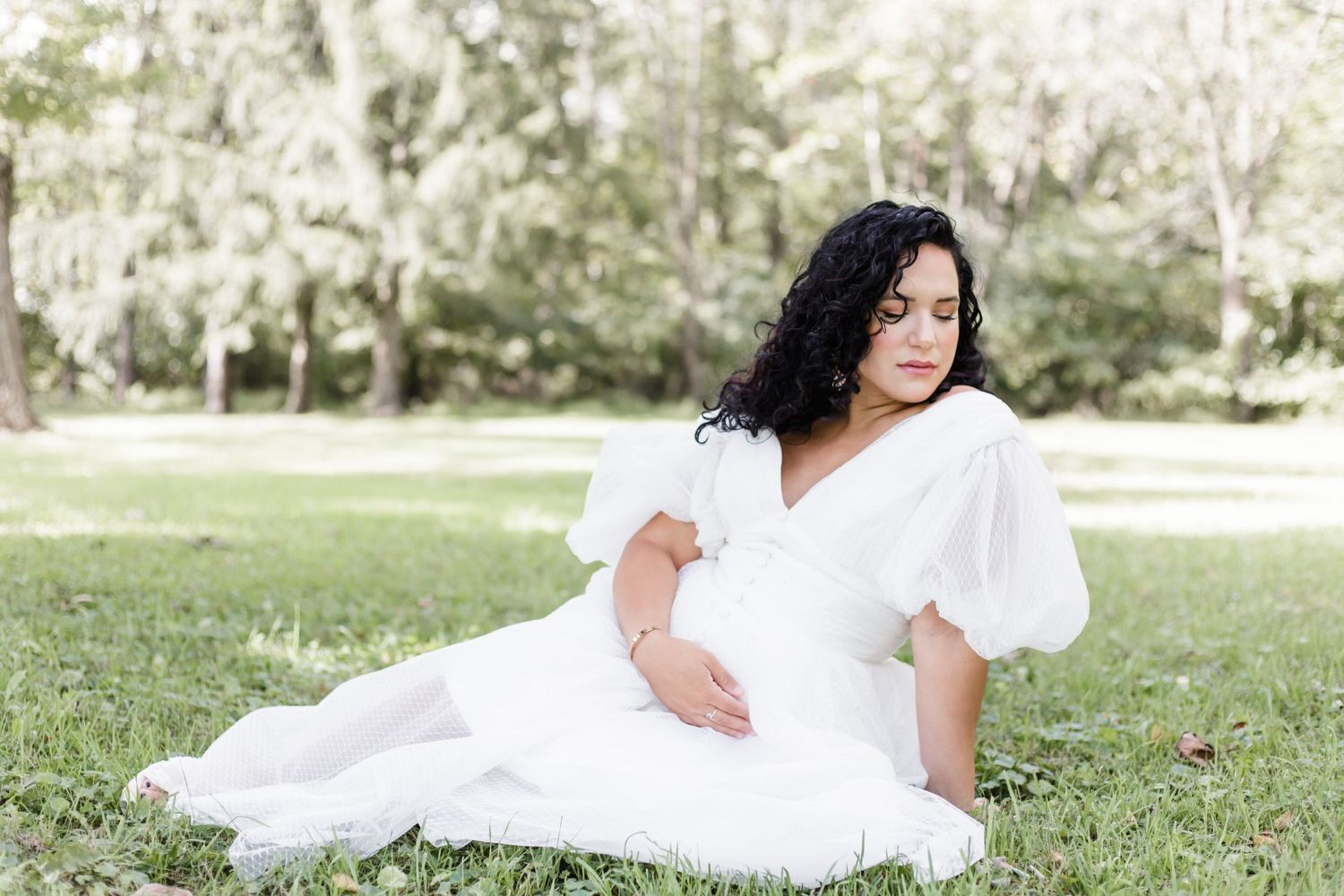 A pregnant mother in a flowing white dress poses gracefully on a grassy lawn surrounded by trees in Lehigh Valley, PA Lock Ridge Furnace.