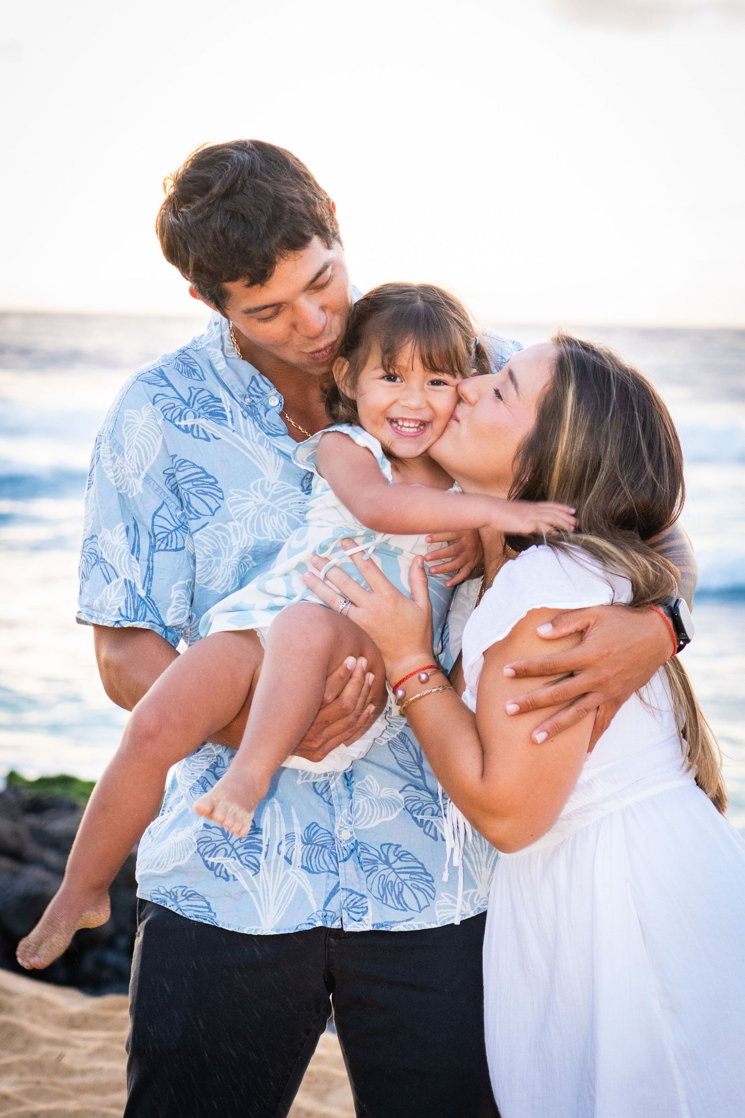 Happy family on a sandy Hawaii beach at sunset, parents kissing their smiling toddler daughter.