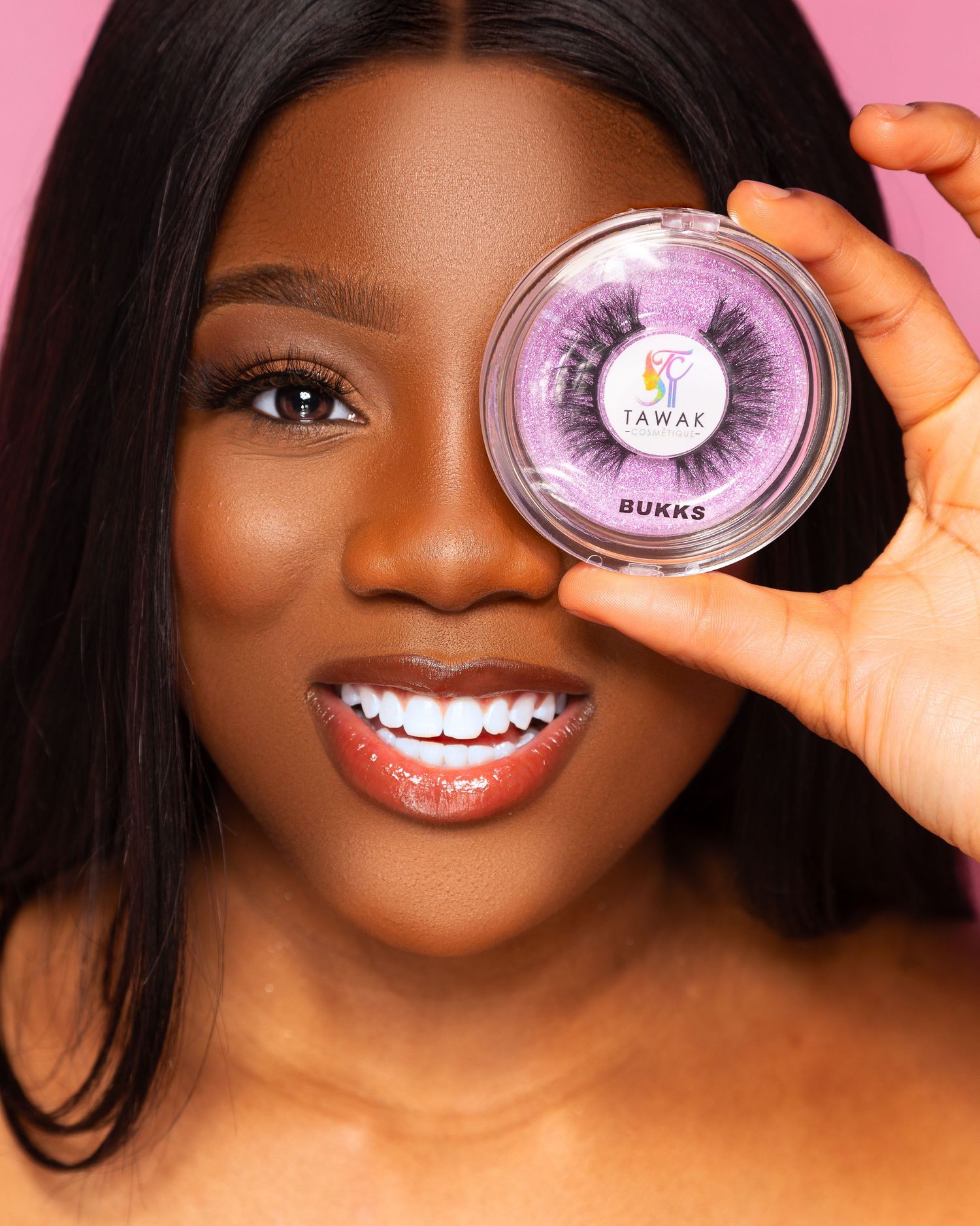 Smiling woman holding Jawak Bukks false lashes container over her eye against pink background.
