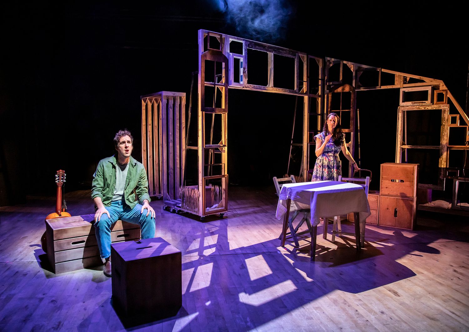 Minimalist theater set with wooden frames and performers on elevated platforms under purple lighting.