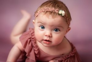 Baby in mauve dress with delicate floral headband lying on pink background.