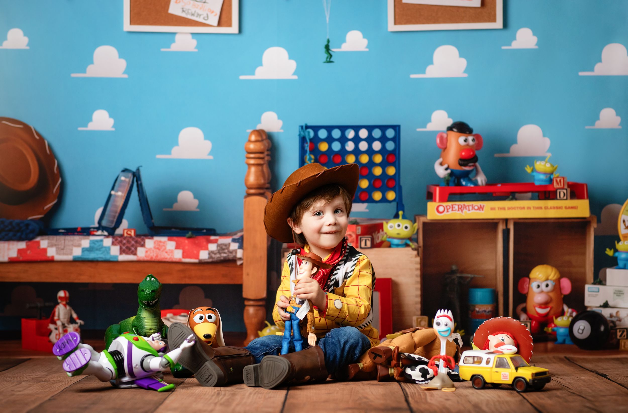 Child dressed as Woody from Toy Story surrounded by character toys in a themed bedroom set with blue sky walls.